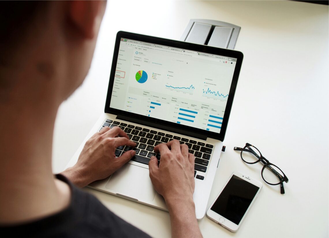 A person analyzing data on a laptop displaying graphs and statistics, with a smartphone and glasses on the desk.