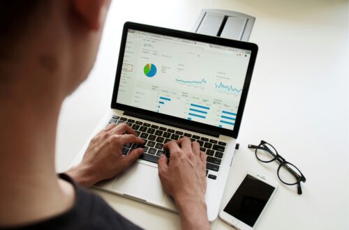 A person analyzing data on a laptop displaying graphs and statistics, with a smartphone and glasses on the desk.